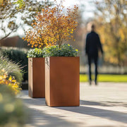 Copper planters with trees and shrubs in an outdoor setting with a blurred person walking in the background.